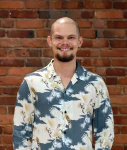 Ryan stands smiling, wearing a patterned shirt in front of a brick background.